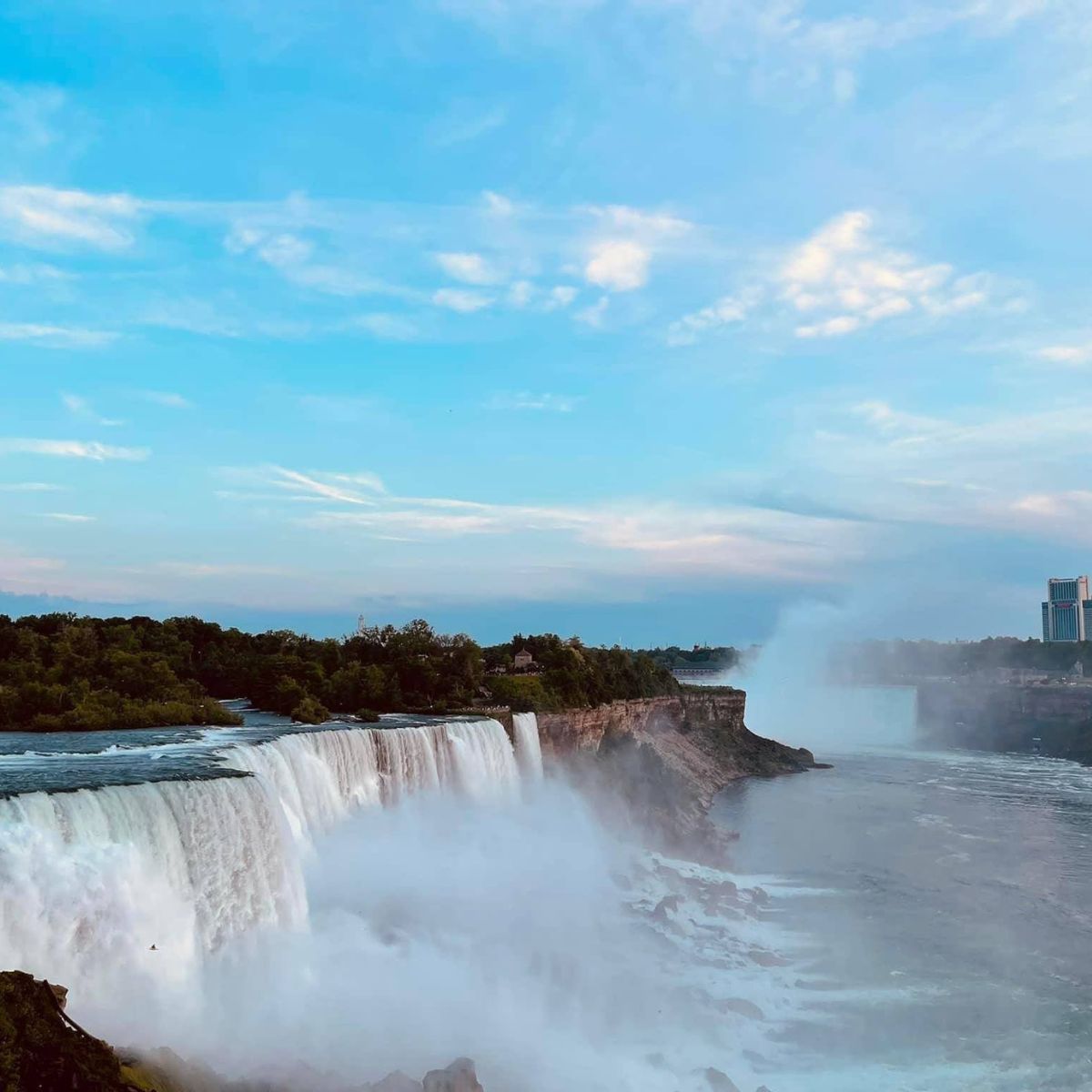 A wide waterfall cascades over a rocky ledge into a misty river under a partly cloudy sky. Trees and a building are visible in the distance, making it one of the perfect day trips from Buffalo.
