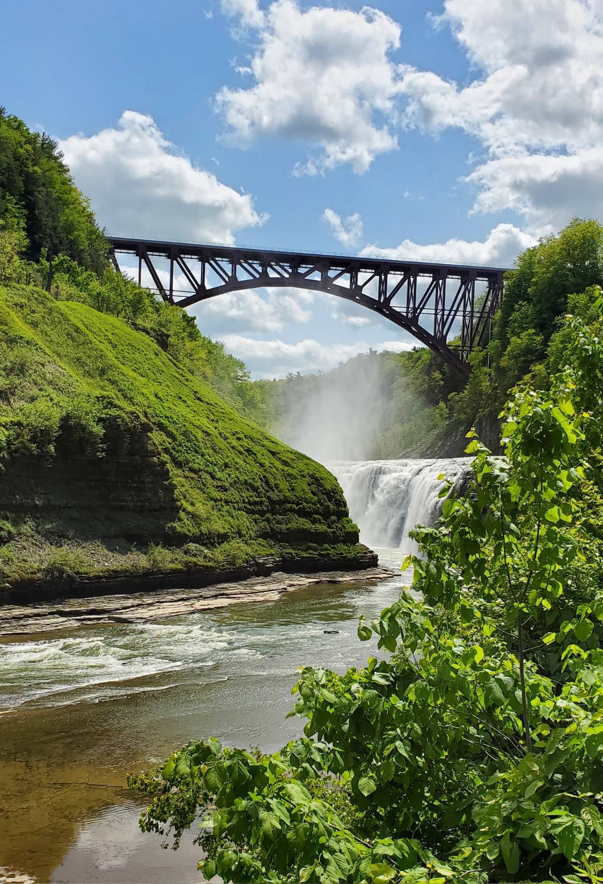 A steel bridge spans over a flowing waterfall surrounded by lush greenery, under a partly cloudy sky.