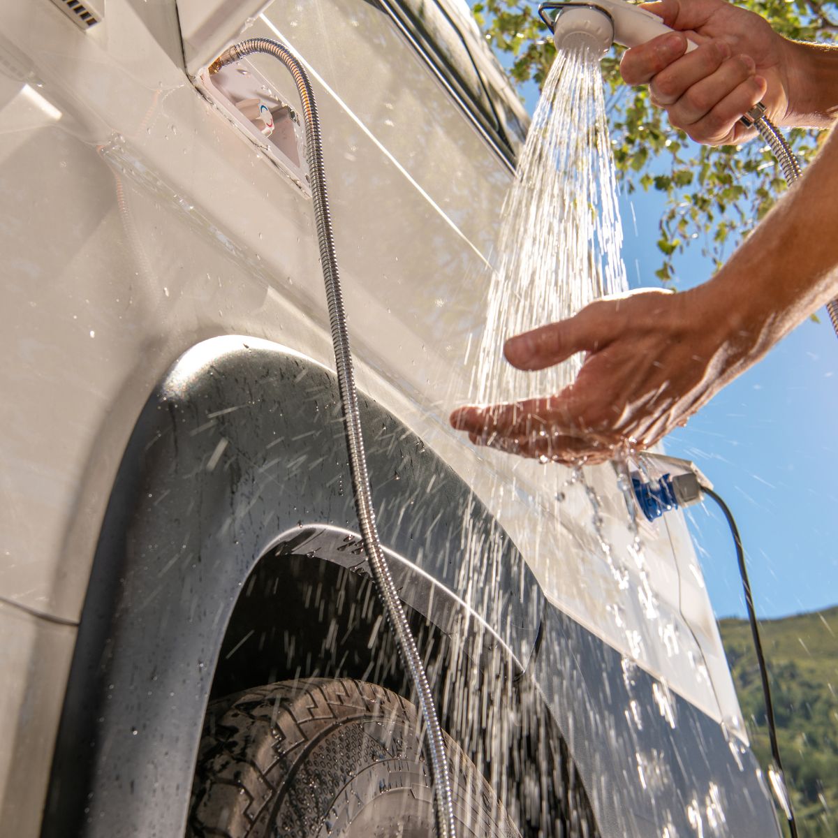 A person uses a handheld showerhead to wash their hands beside a white vehicle, outdoors.