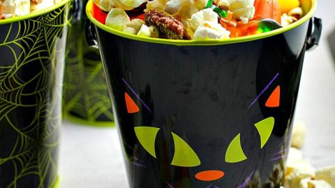 A black bucket with a Halloween face design filled with popcorn, candy corn, and nuts. Other similar buckets are partially visible in the background.
