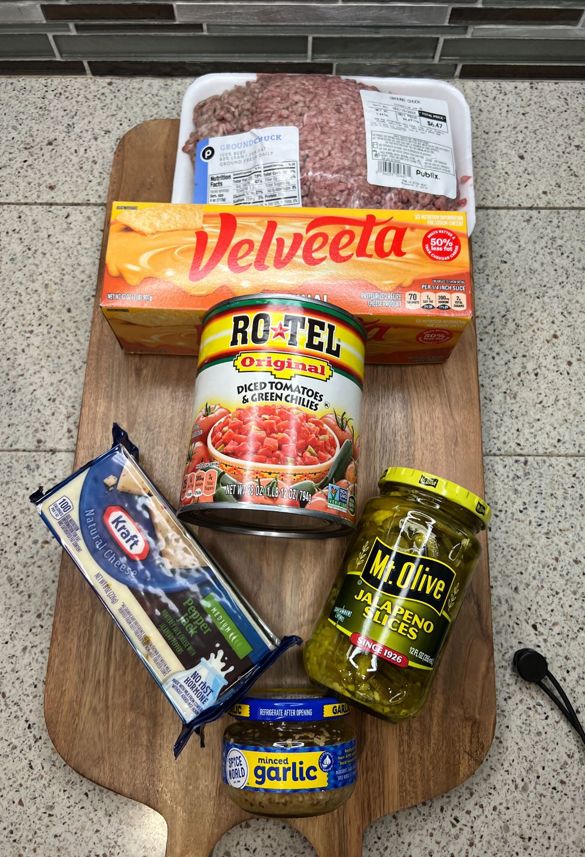 Various food items, including ground beef, Velveeta cheese, diced tomatoes with green chilies, jalapeño slices, minced garlic, and a pack of cheddar cheese, displayed on a wooden board.
