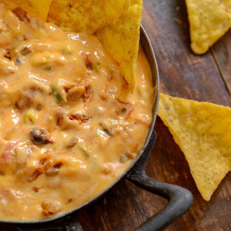 A skillet of cheesy smoked queso dip with visible chunks of meat and peppers, surrounded by tortilla chips on a wooden surface.