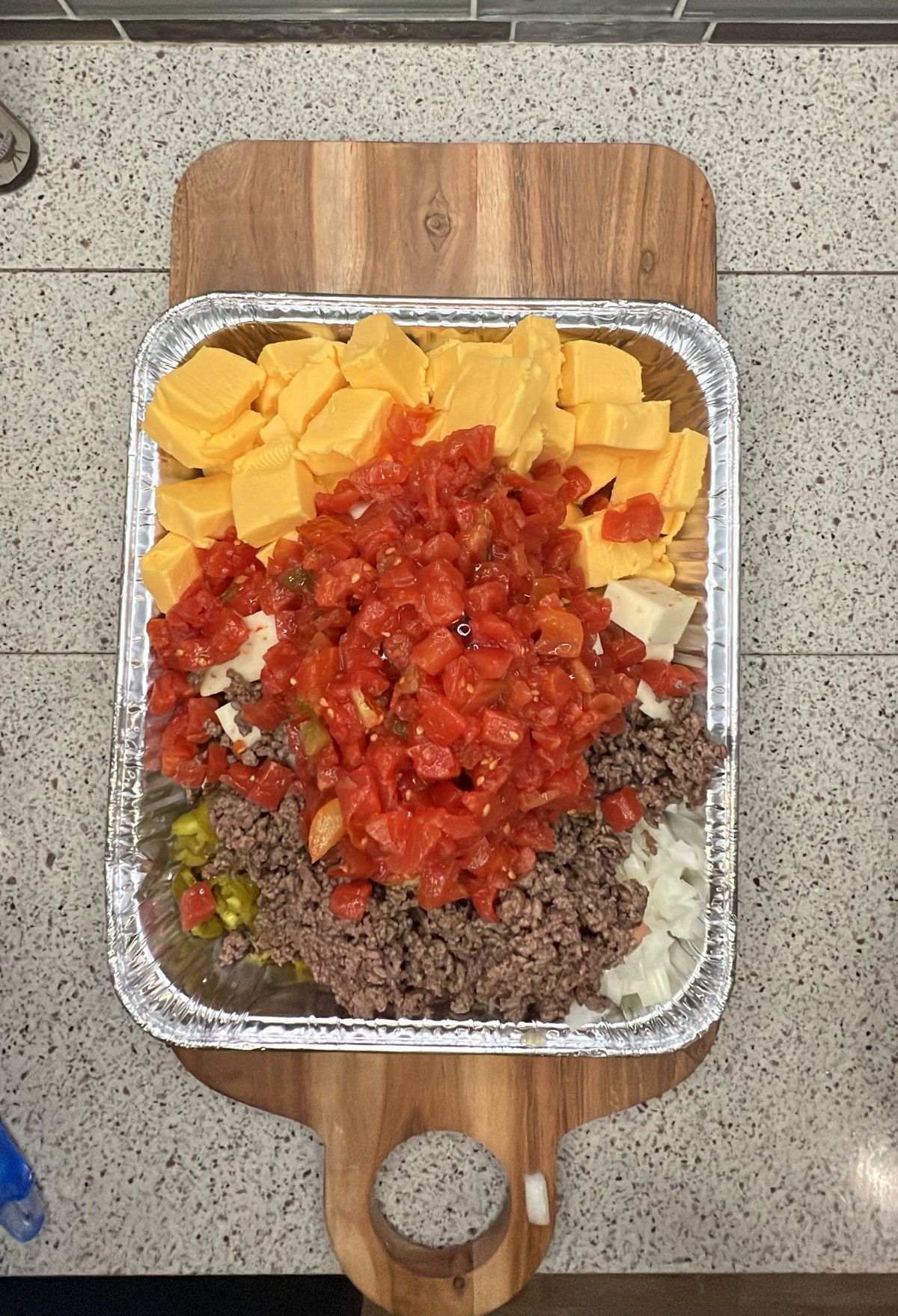Metal tray on a wooden board containing diced cheese, ground beef, chopped tomatoes, jalapeños, and onions, ready for mixing or cooking.