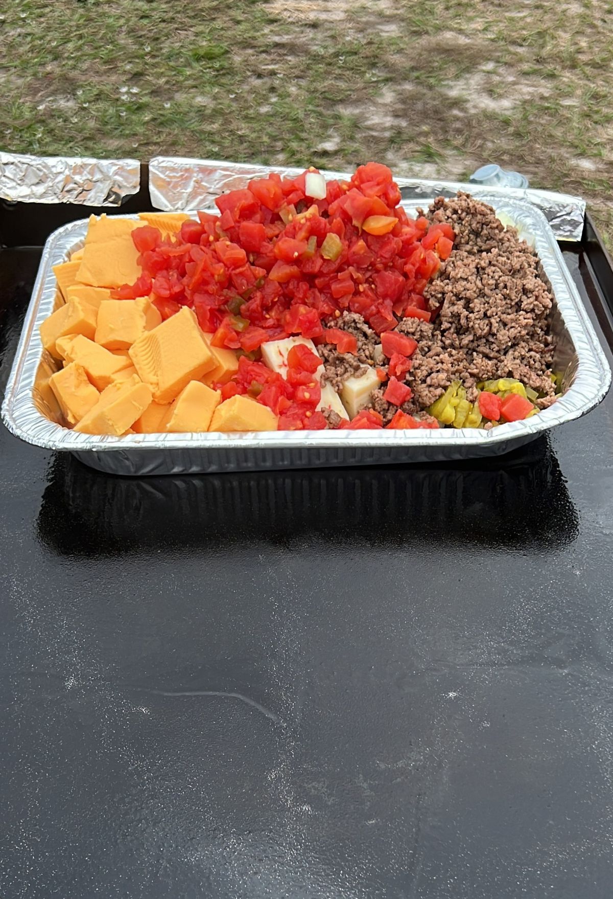 A foil tray filled with cubed cheese, ground beef, diced tomatoes, and peppers on a black surface outdoors.