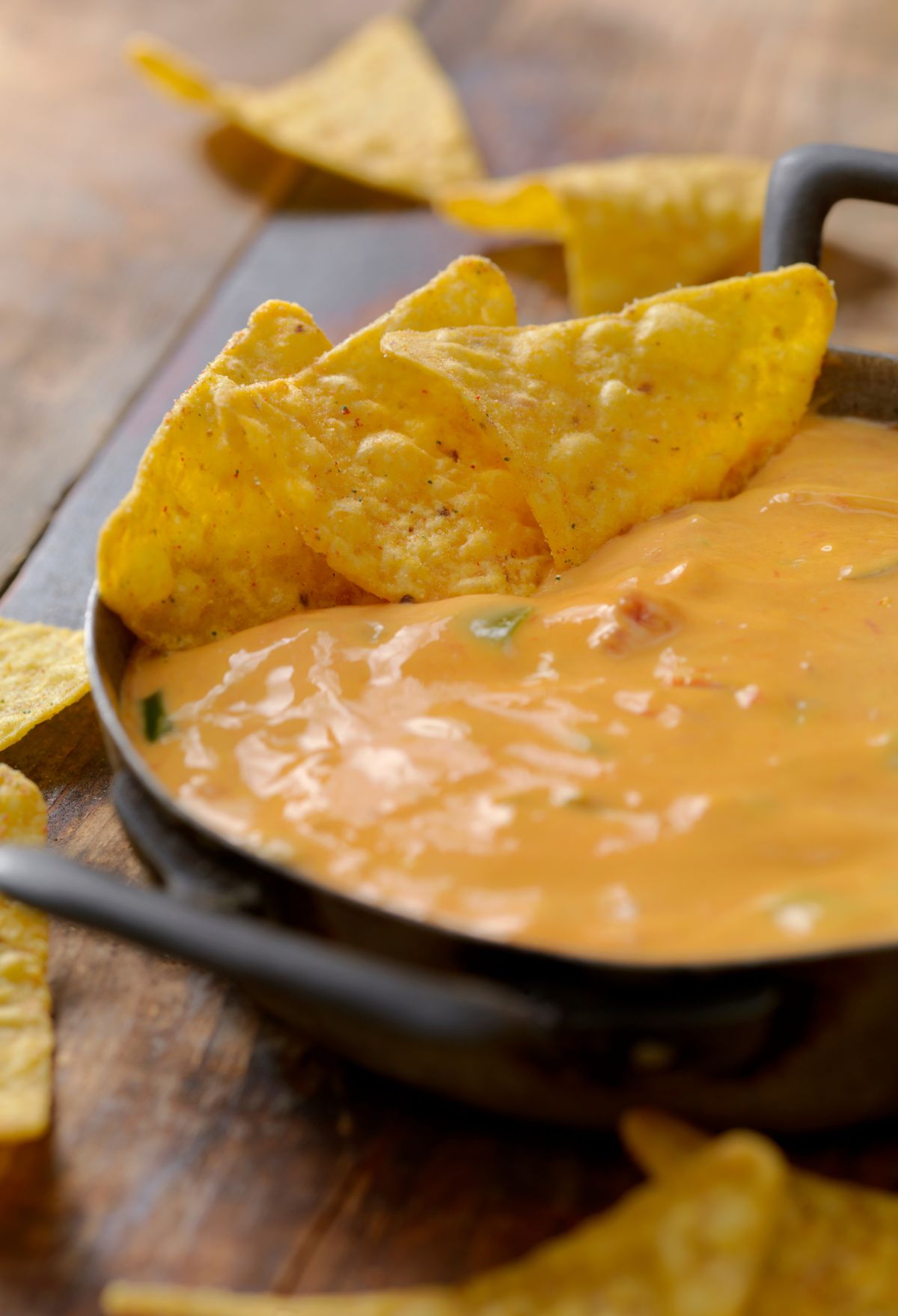 A bowl of creamy cheese dip with pieces of green herbs and tomatoes, accompanied by tortilla chips on a wooden table.