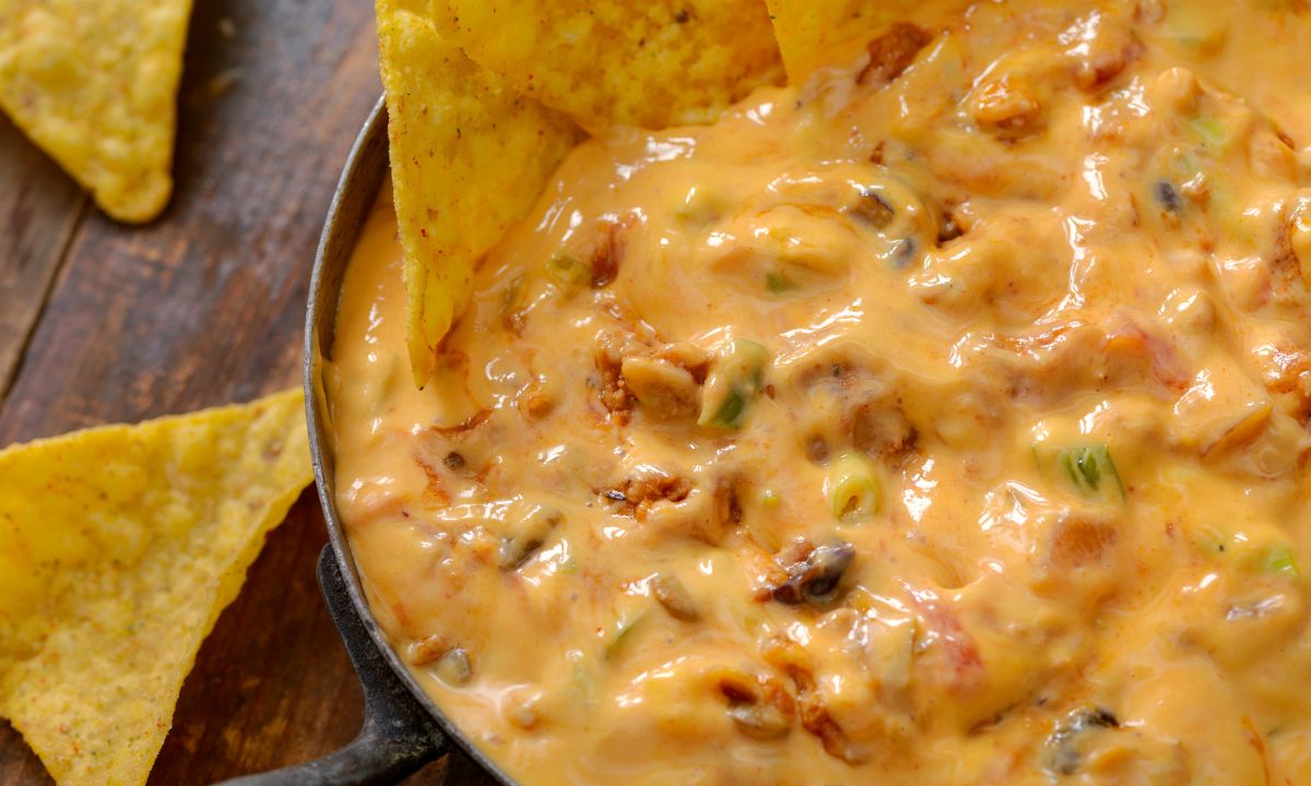 Close-up of a skillet filled with creamy cheese dip containing pieces of meat and vegetables, surrounded by tortilla chips on a wooden surface.