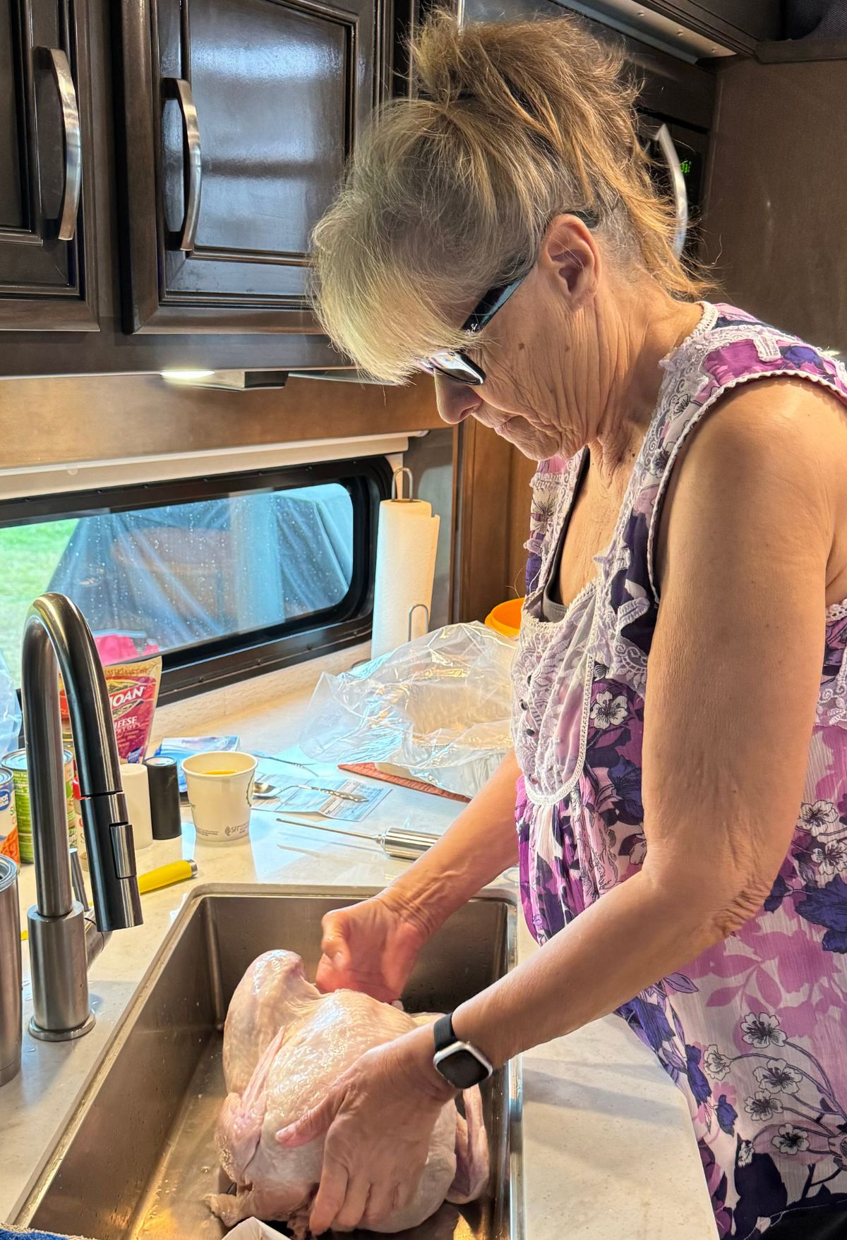 A woman in a floral apron and glasses washes a raw turkey by the kitchen sink, preparing it for the convection oven. Kitchen items and a window provide a cozy backdrop.