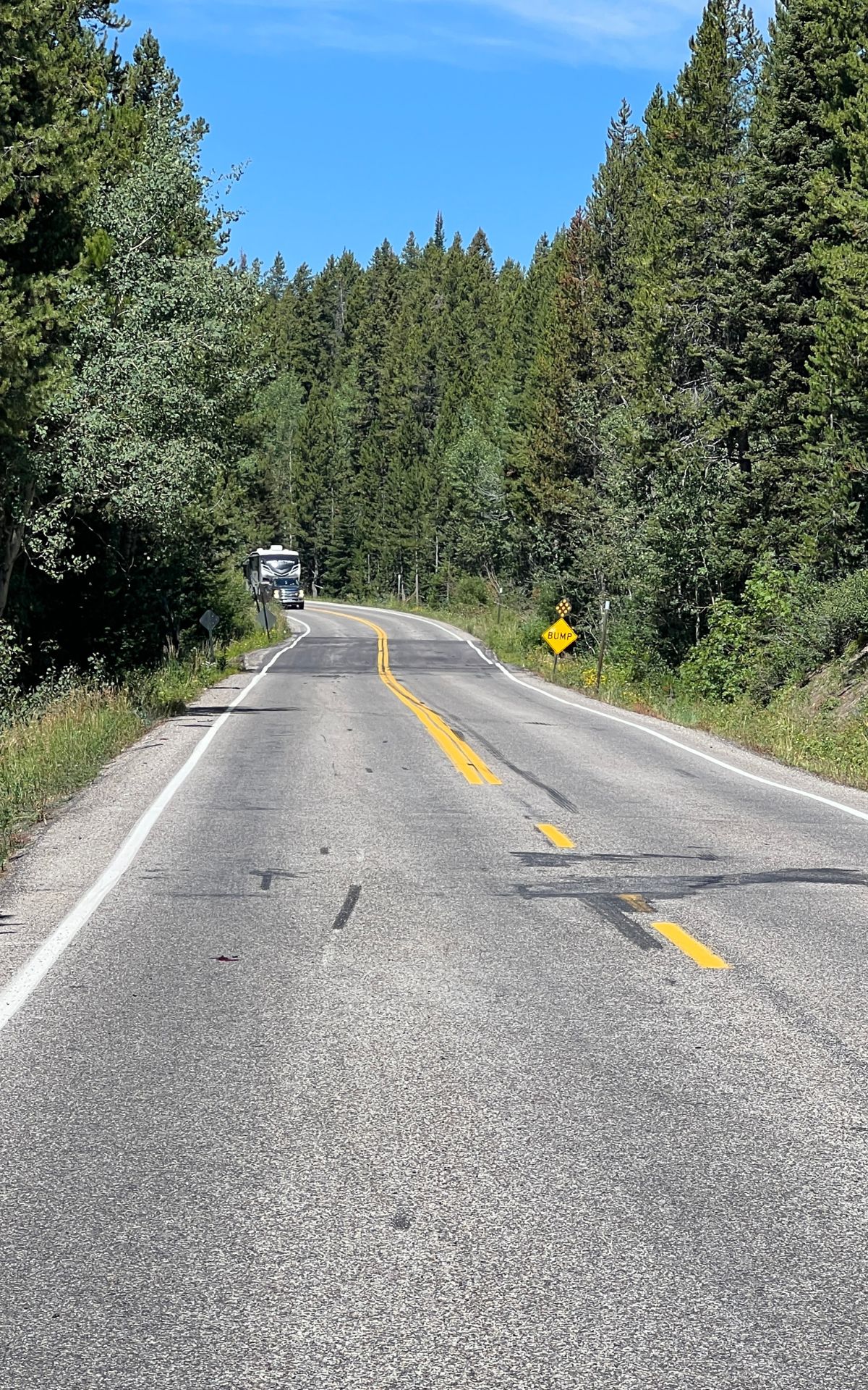 A two-lane road curves through a forested area, with a truck in the distance and a yellow caution sign on the right side—perfect scenery for those who love to say, “Lets RV.”.