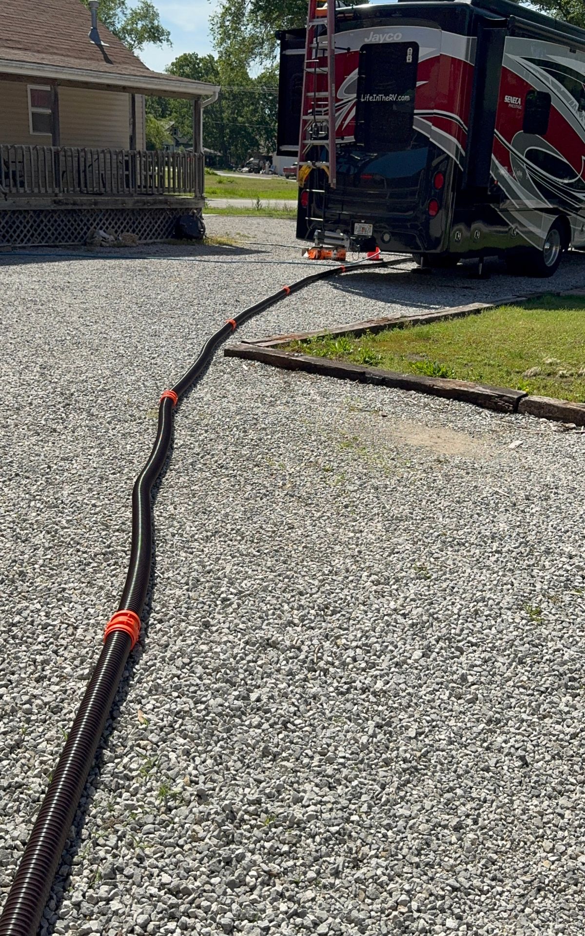 A long black hose, secured with orange brackets, connects to the back of an RV on a gravel driveway—just another day living full-time in an RV.