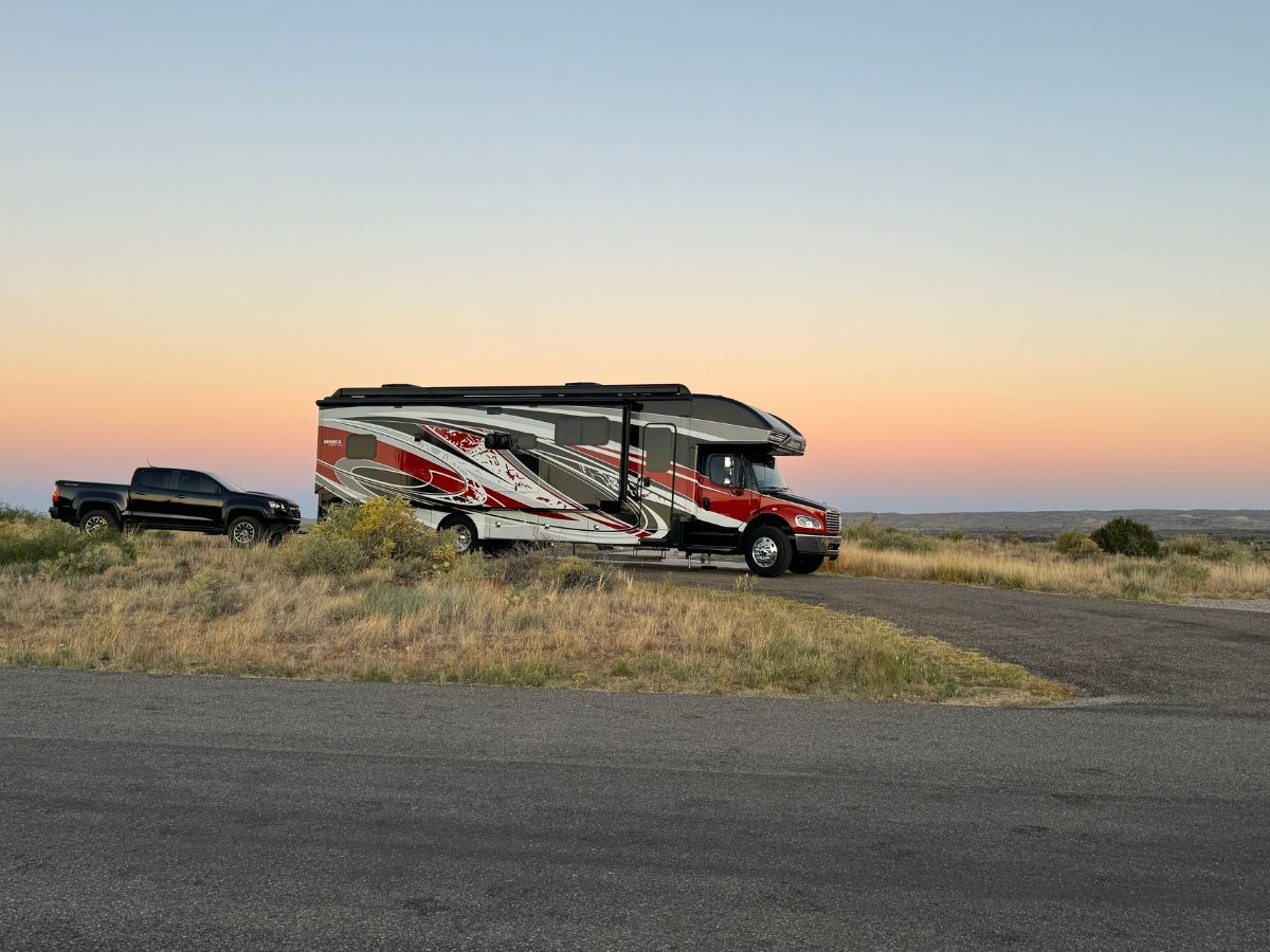 A black and red RV, possibly serving as a new home, is parked on a paved area beside a pickup truck in an open, grassy landscape at sunset.