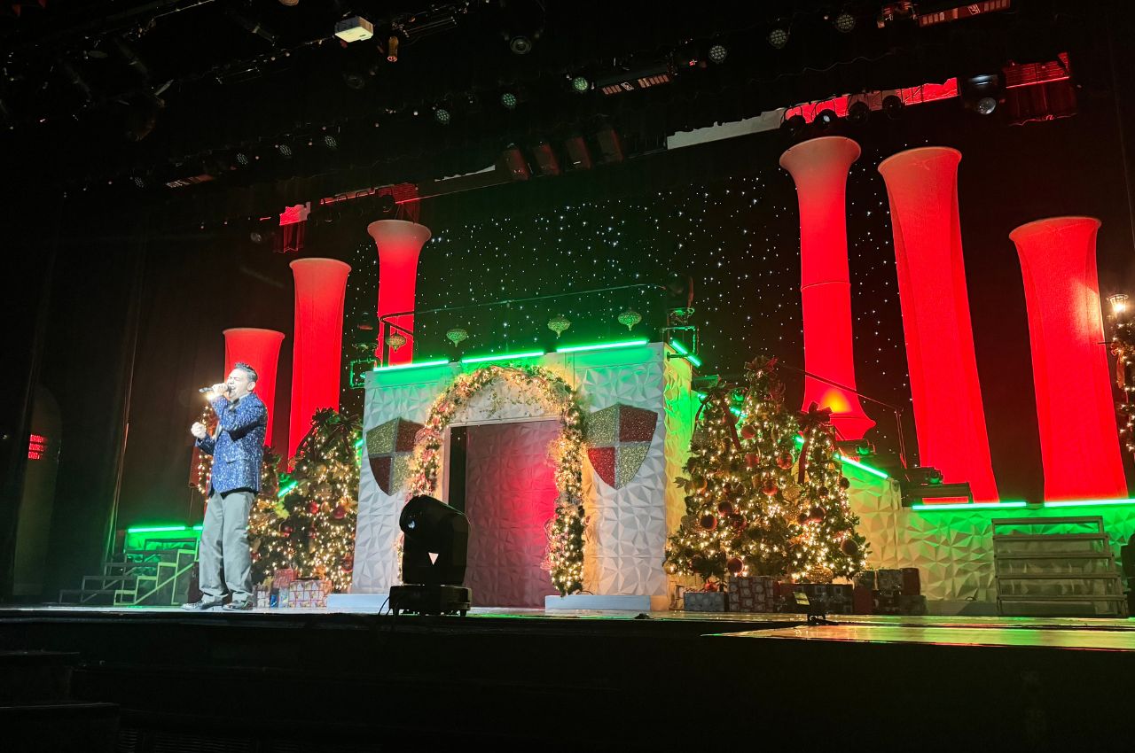 A performer stands on stage next to holiday decorations, including Christmas trees, presents, and columns illuminated with red and green lights at Branson Christmas Wonderland.