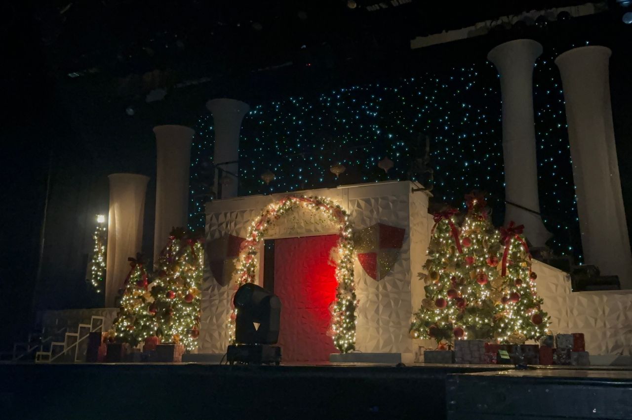 A stage decorated with white columns, Christmas trees, garlands with lights, wrapped presents, and a red door sets the festive scene for Branson Christmas Wonderland against a backdrop of blue twinkling lights.