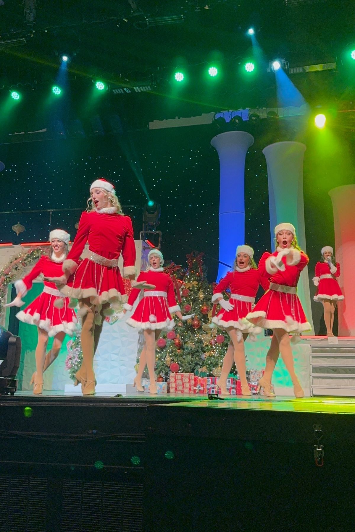 Performers in matching red and white holiday costumes dance on stage in Branson Christmas Wonderland, surrounded by Christmas trees, gifts, and festive decorations under colorful stage lights.
