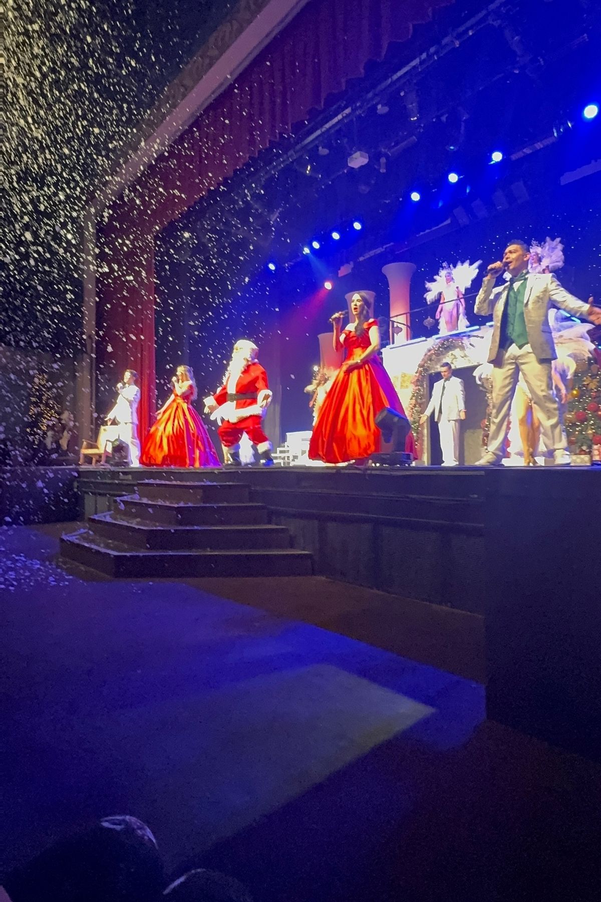 Performers in formal attire and a Santa Claus costume stand on a decorated stage at Branson Christmas Wonderland, with artificial snow falling and blue stage lights illuminating the festive scene.
