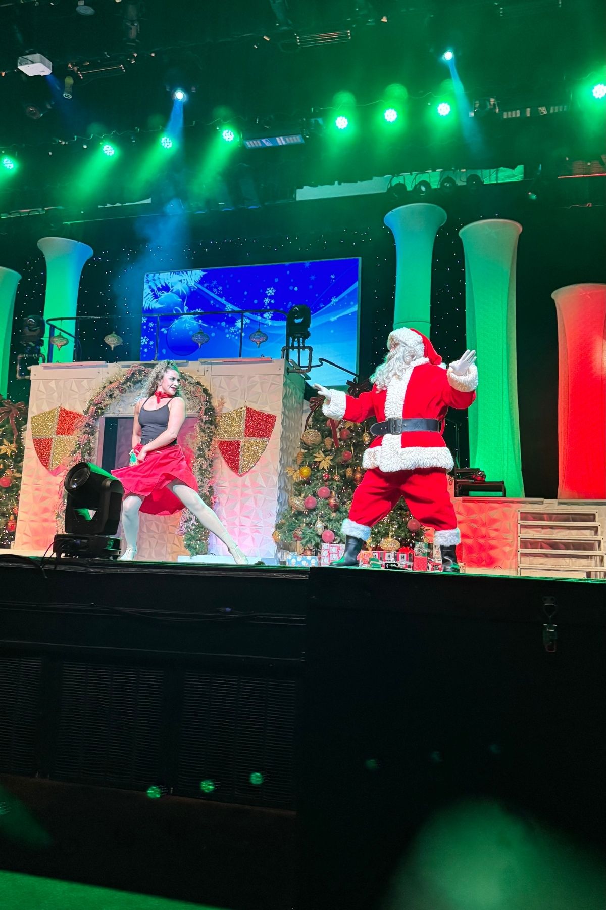 A performer in a red dress dances next to a person dressed as Santa Claus on a festively decorated Branson Christmas Wonderland stage with green and red lighting.