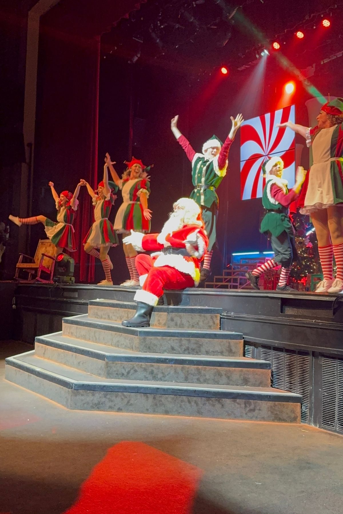 Santa Claus sits on steps in front of a festive Branson Christmas Wonderland stage while performers dressed as elves and candy canes dance in the background under red lights.