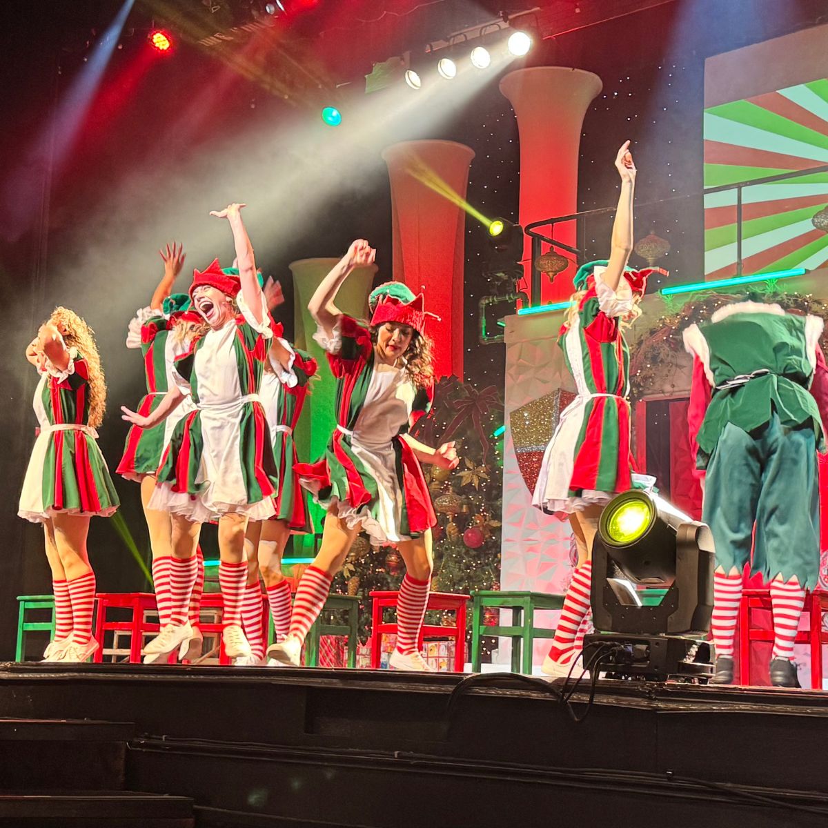 A group of performers dressed as Christmas elves dance on stage under colorful lighting at Branson Christmas Wonderland, wearing red and green costumes with striped socks.