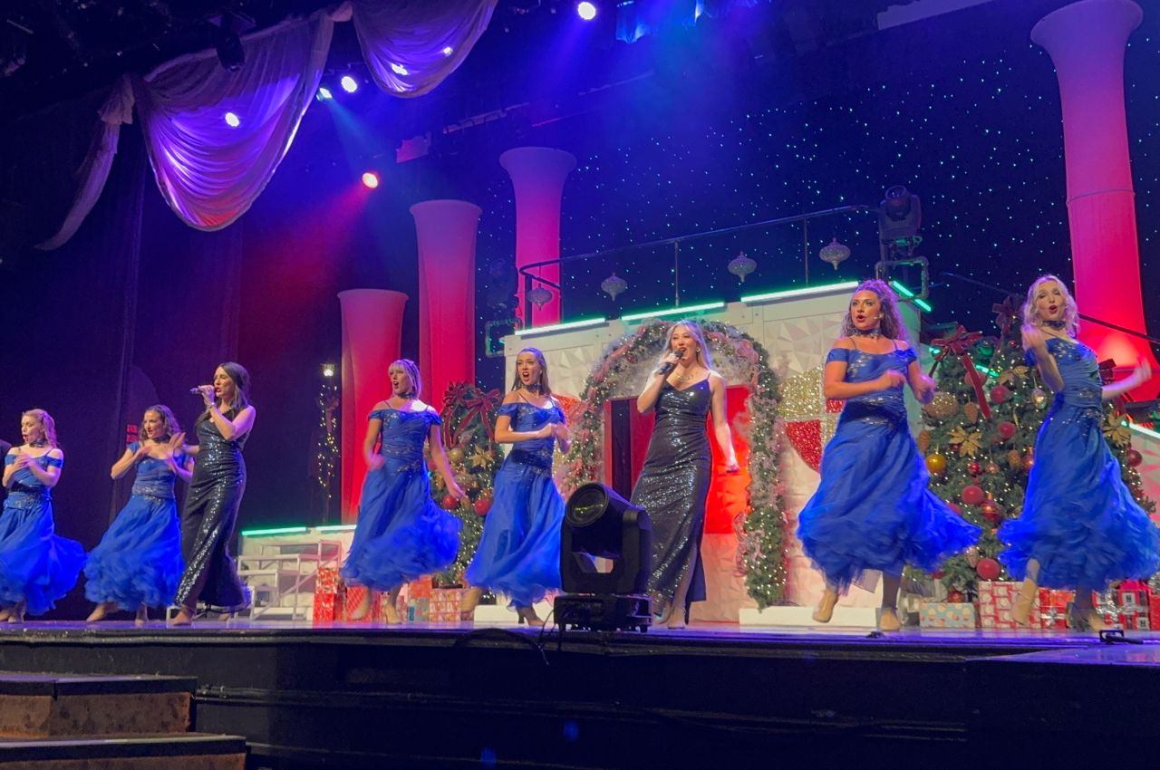 A group of women in blue dresses perform a choreographed dance and vocal number on the brightly lit, decorated stage of Branson Christmas Wonderland, complete with festive columns and holiday decorations.