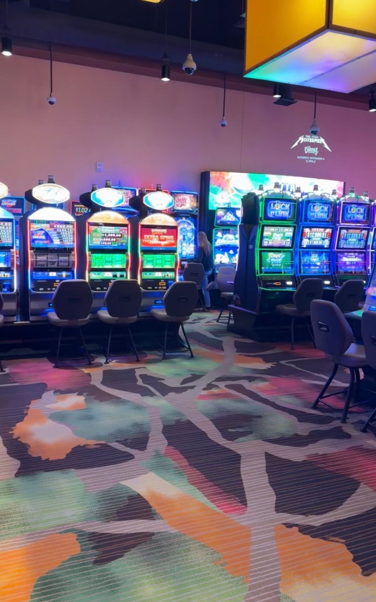 A row of colorful slot machines in a casino sits empty, chairs lined up as if awaiting guests on a casino camping adventure, with patterned carpet stretching beneath them.