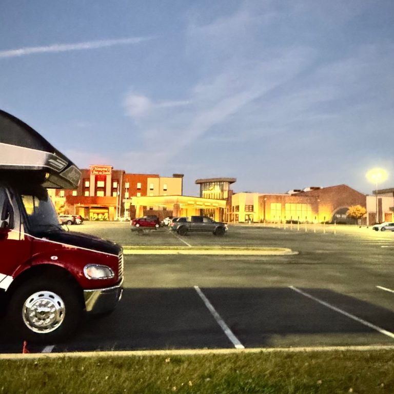 A nearly empty parking lot at dusk features a red vehicle in the foreground and a well-lit casino in the background, offering a perfect spot for casino camping enthusiasts.