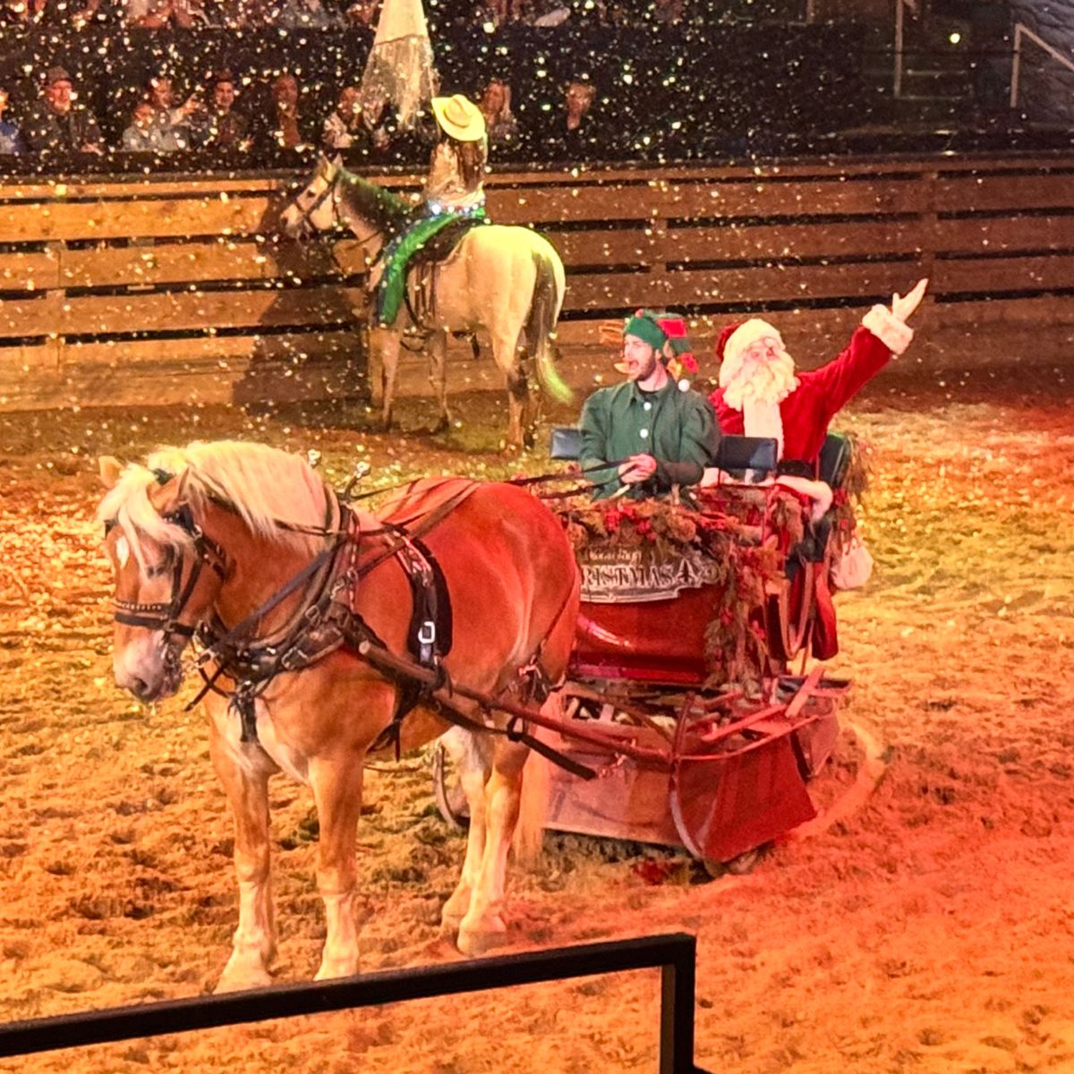 Santa Claus and an elf sit on a holiday-themed wagon pulled by a horse inside an arena, with artificial snow falling around them as the RV Christmas Trail audience watches in delight.