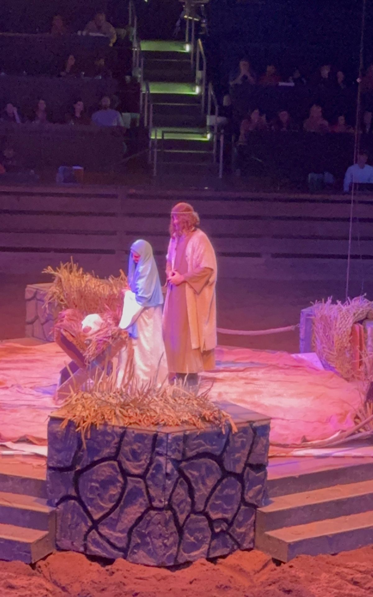 Two people in biblical costumes stand beside a manger on a circular stage during the RV Christmas Trail, with an audience seated in the background.