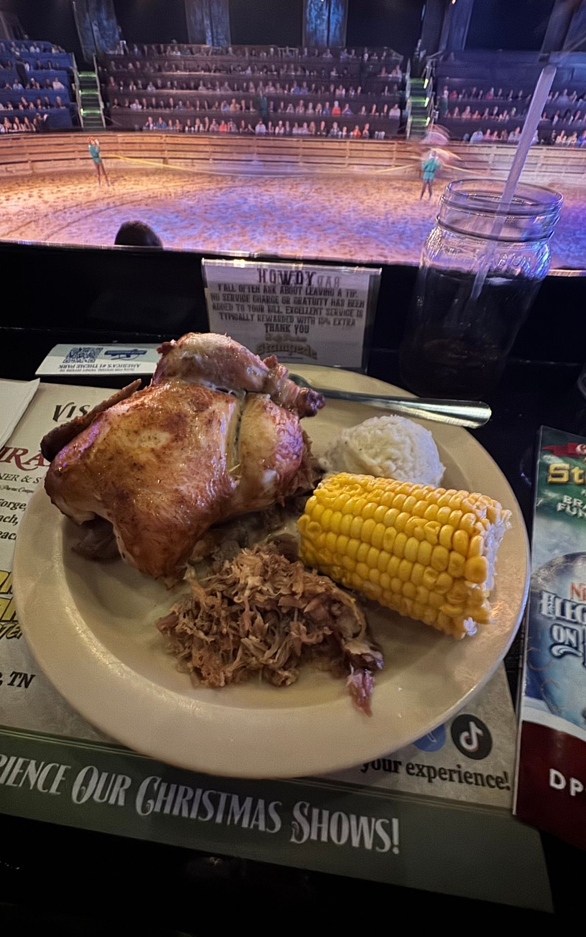 A plate with roast chicken, pulled pork, corn on the cob, and mashed potatoes sits on a table beside a drink and papers, capturing the cozy atmosphere of the RV Christmas Trail with a show arena in the background.