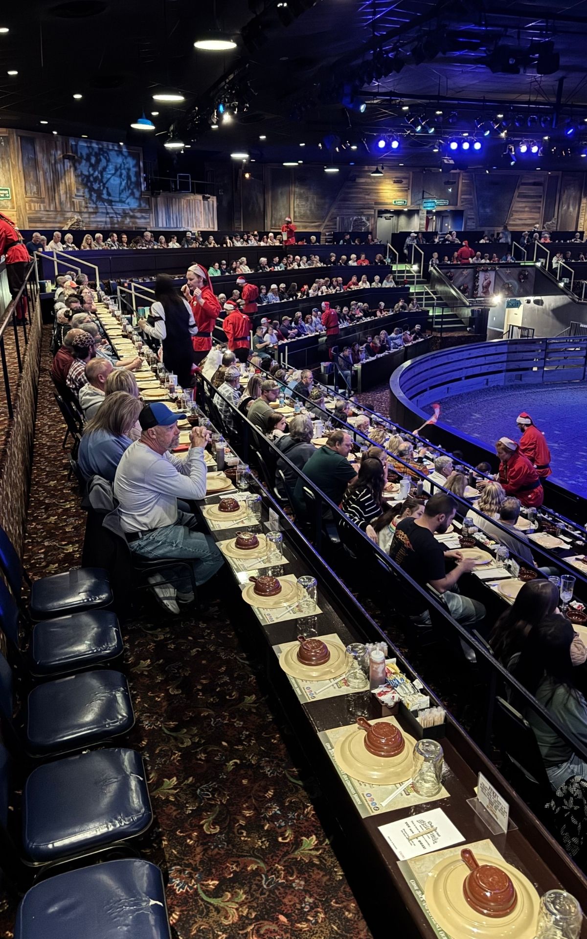 Audience members sit at long tables set with plates and mugs, waiting for a show to begin in a large indoor theater with tiered seating and an arena-style stage—perfect for those enjoying the holiday magic of the RV Christmas Trail.