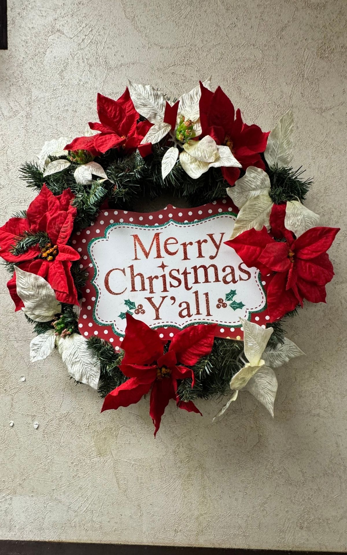 A Christmas wreath with red and white poinsettias surrounds a sign that reads "Merry Christmas Y'all," welcoming visitors on the festive RV Christmas Trail.