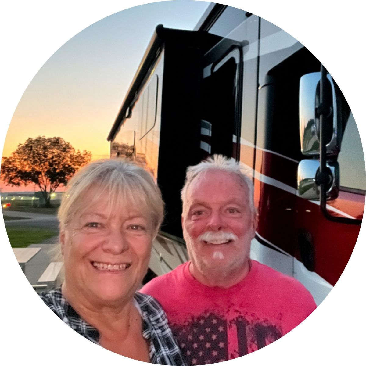 Two smiling adults stand in front of a large RV at sunset, with trees and a road visible in the background.