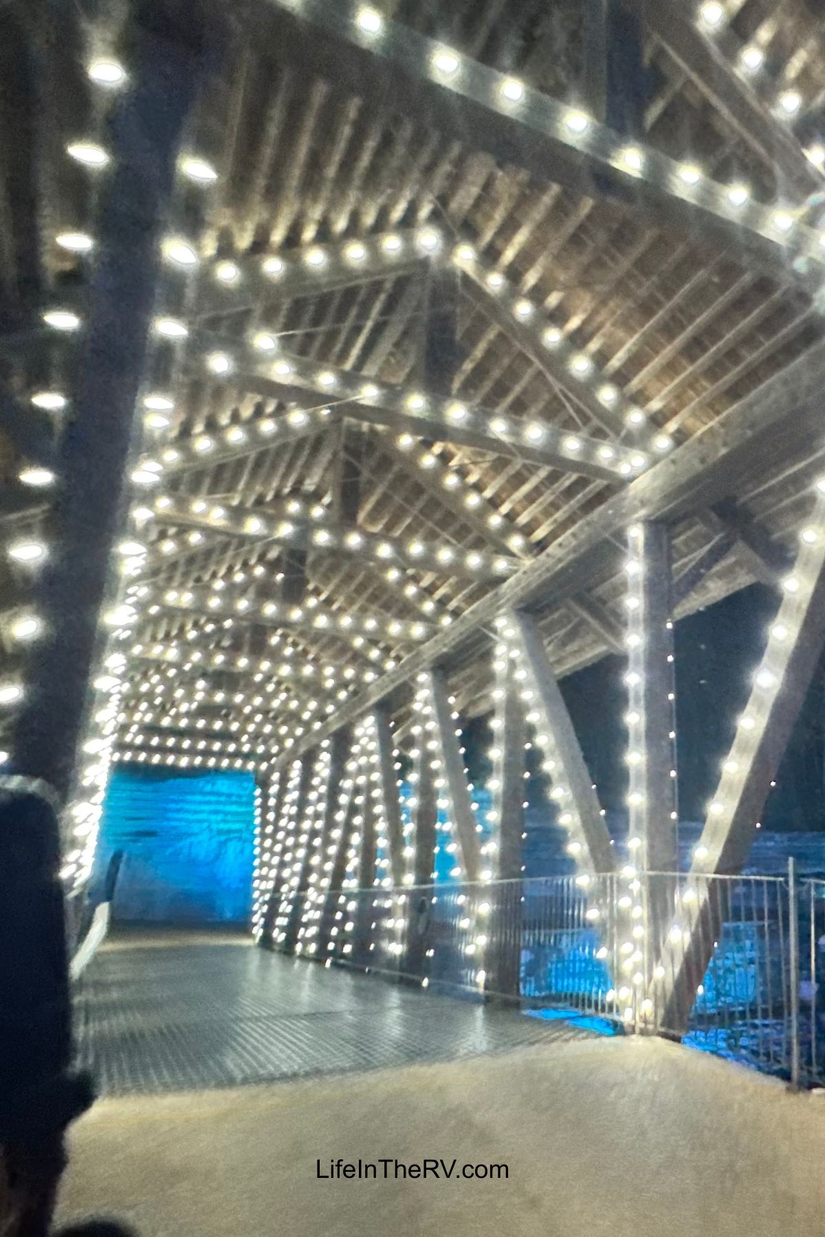 A wooden bridge, popular among Holiday RV Destinations, is decorated with strings of white lights and illuminated in blue at night.