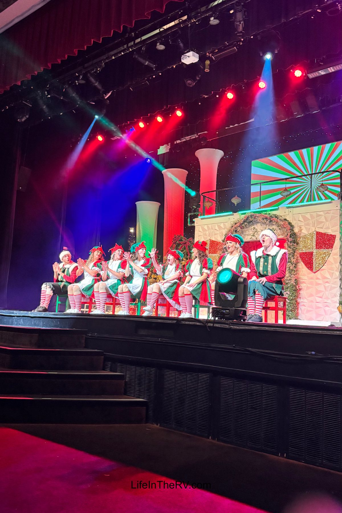Performers dressed as elves sit on stage under colorful lights, participating in a holiday-themed show with festive decorations in the background—perfect inspiration for your next Holiday RV Destinations adventure.