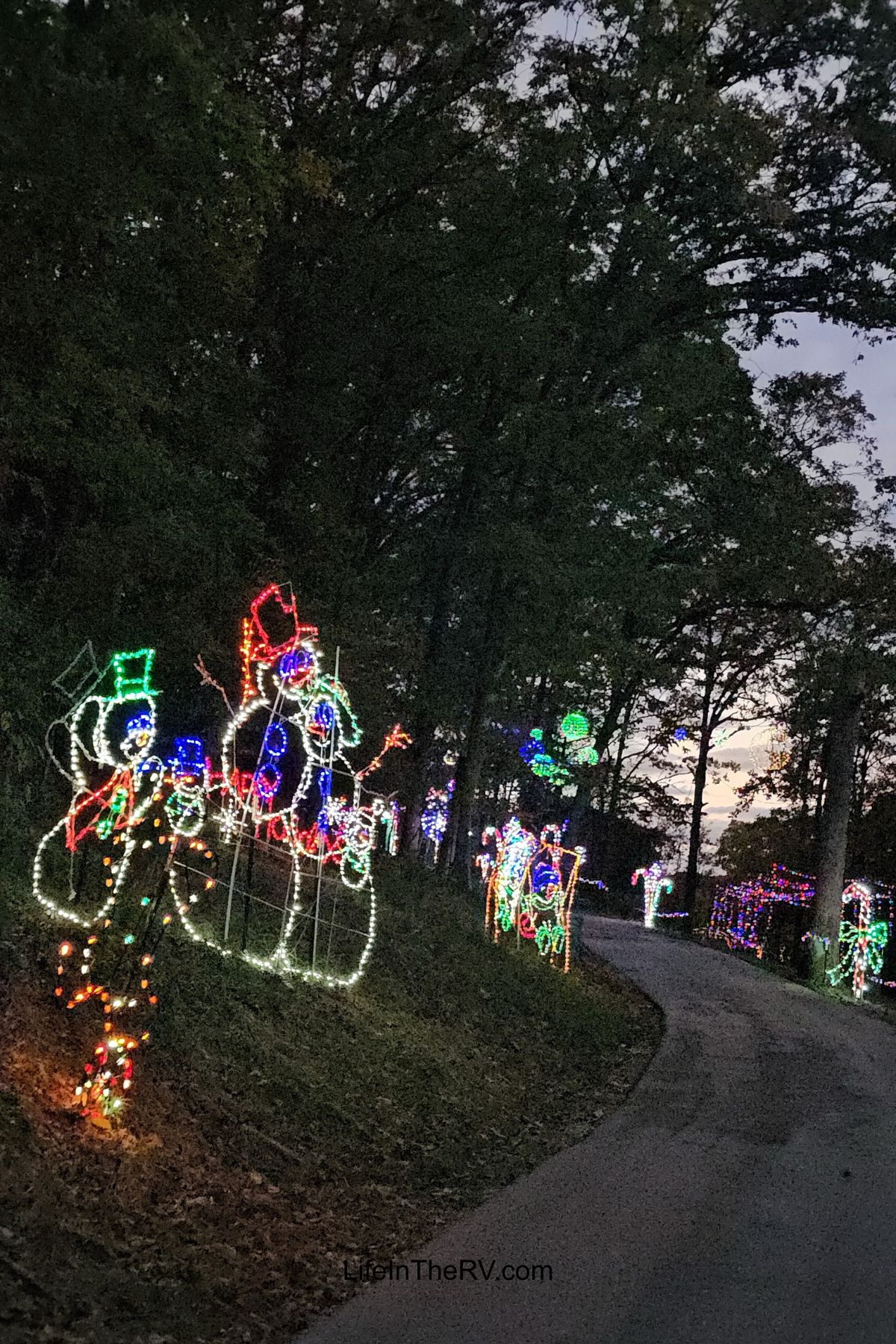 Colorful holiday light displays shaped like snowmen and festive figures line a winding road through a wooded area at dusk, creating the perfect scene for memorable Holiday RV Destinations.