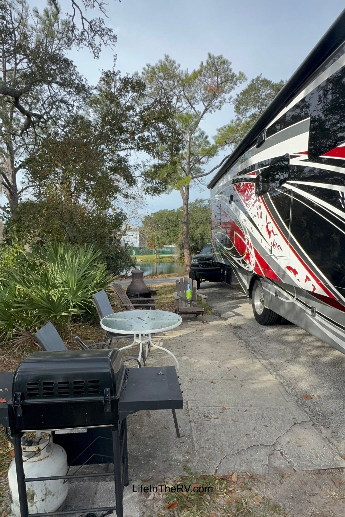 A parked RV next to a patio area with a round table, two chairs, a grill, a propane tank, and a fire pit surrounded by trees and plants—perfect for enjoying top Holiday RV Destinations in comfort and style.