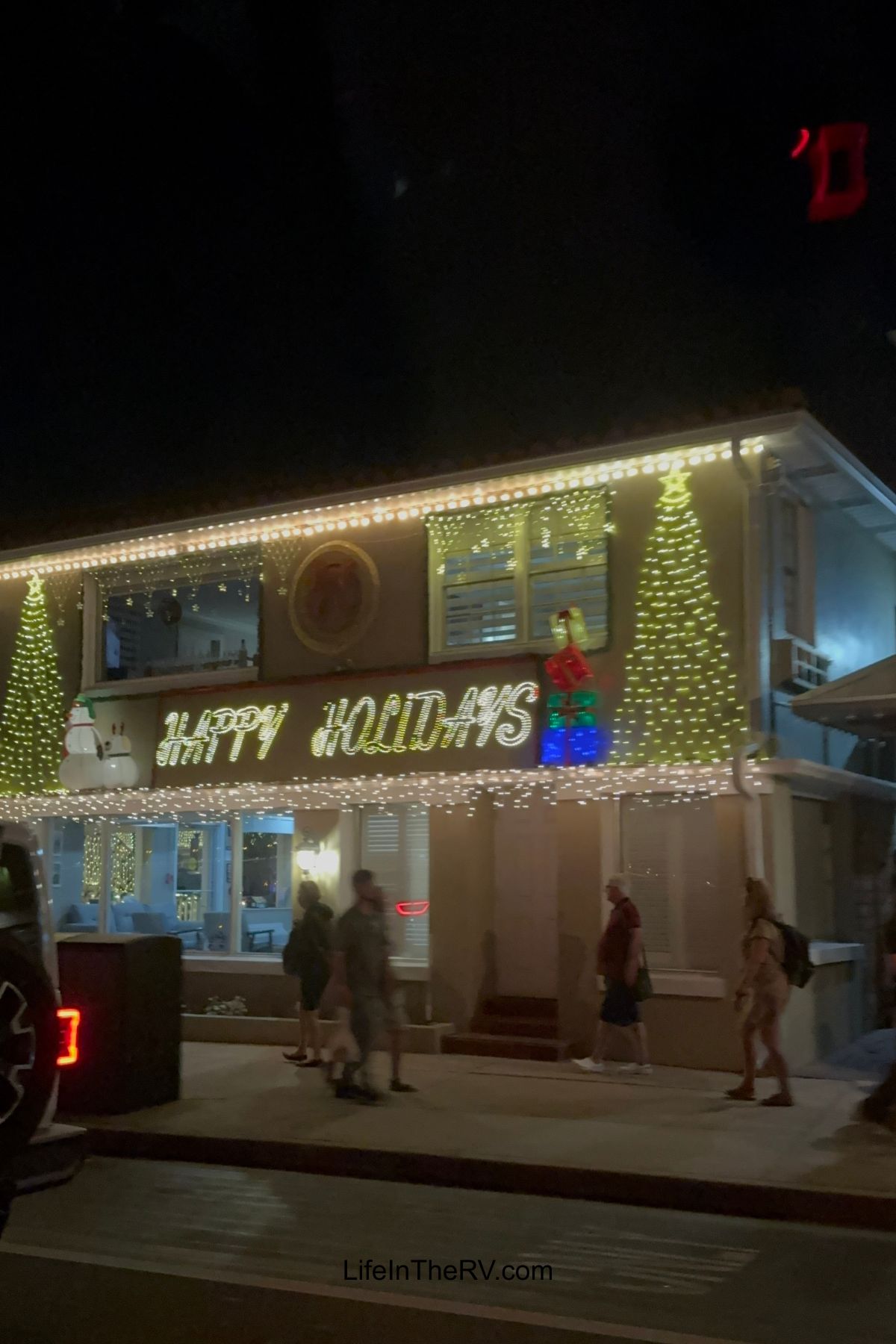 Two-story building decorated with holiday lights, a snowman, and the words "Happy Holidays" on the front, capturing the festive spirit of Holiday RV Destinations. Several people walk by on the sidewalk at night.