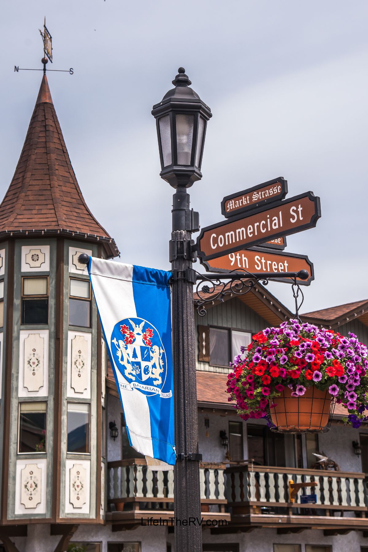 Street sign at the intersection of Commercial Street and 9th Street with a German-style building, a blue and white flag, and a hanging basket of pink flowers—perfect for those exploring unique Holiday RV Destinations.