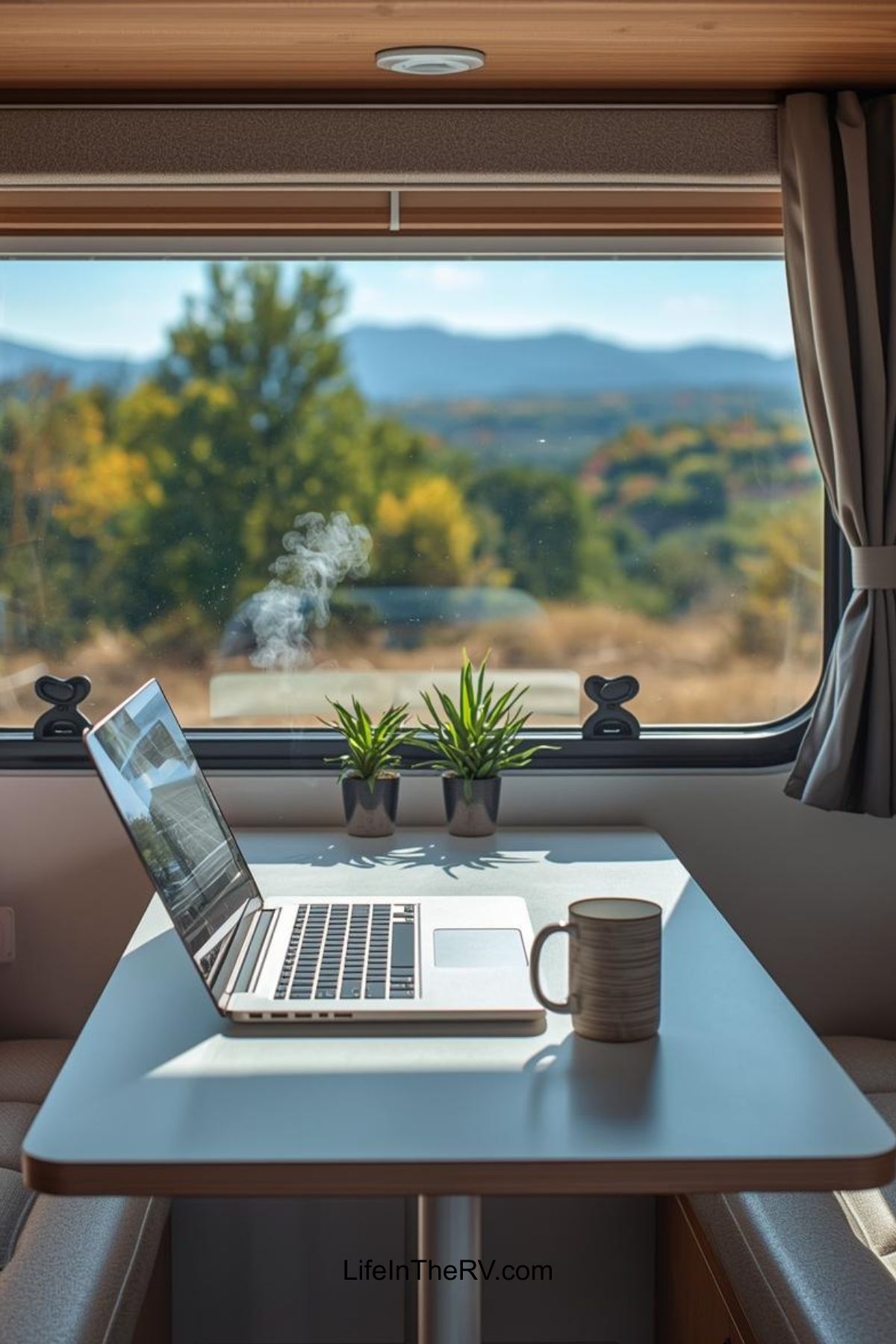 A laptop, a steaming mug, and two small potted plants sit on an RV table by a window with a scenic view—perfect inspiration for learning how to RV full-time amid trees and distant mountains.
