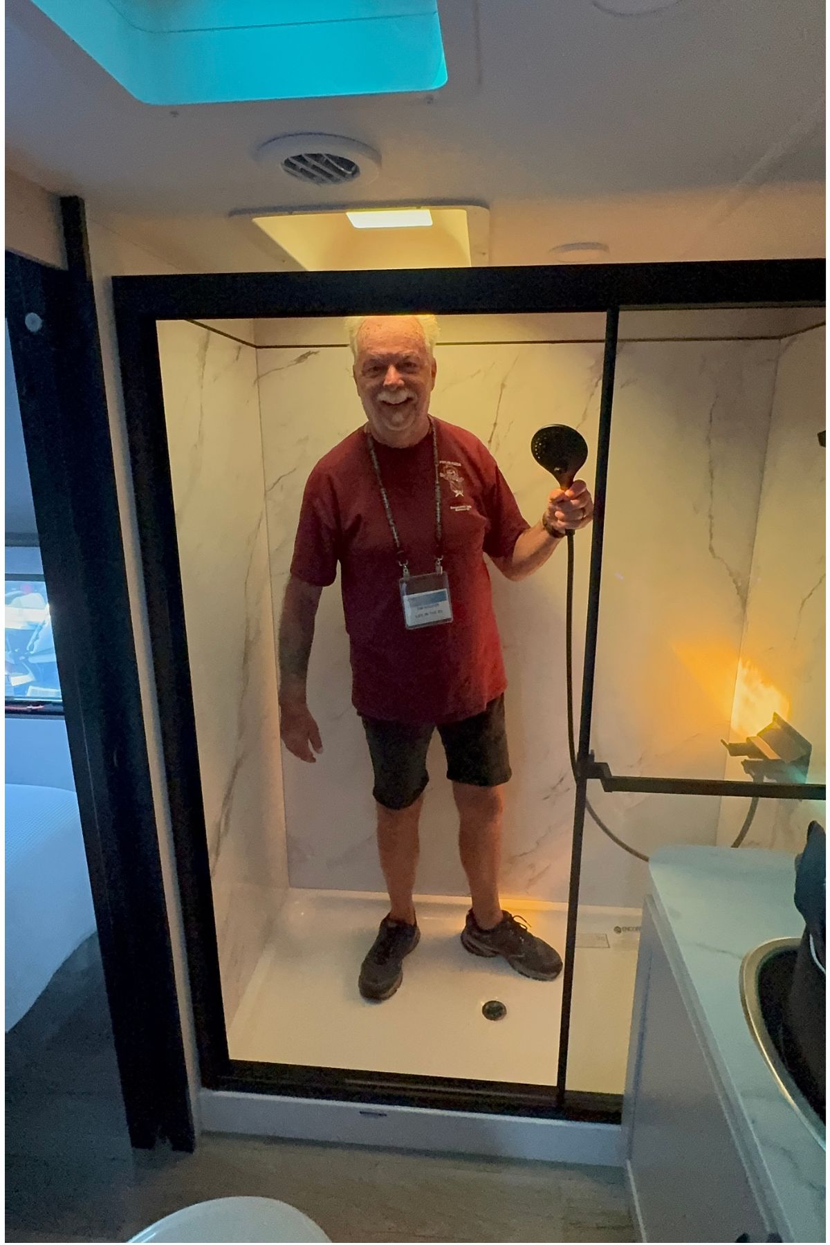 An older man in a maroon shirt and shorts stands smiling in a shower stall, holding a showerhead inside what appears to be an RV bathroom—a glimpse into how to RV full-time with comfort and convenience.