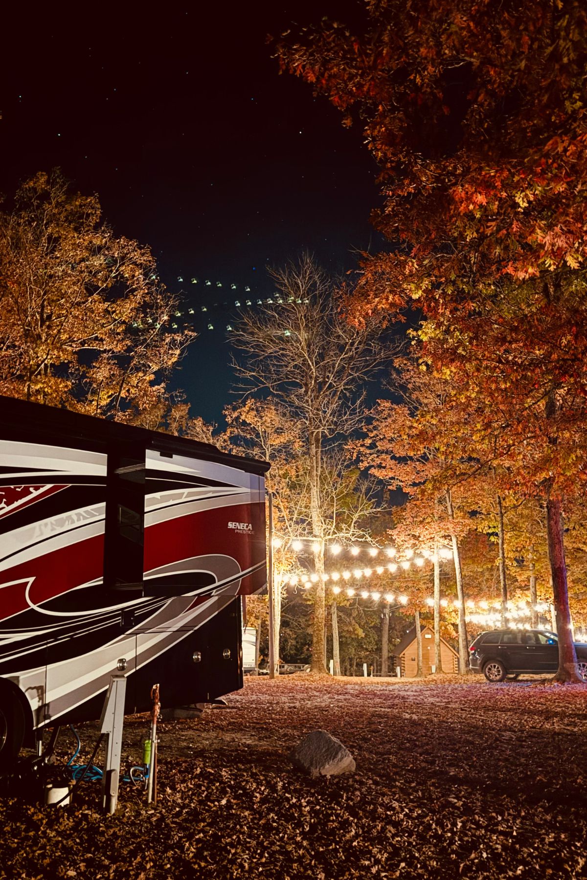 A camper and car are parked among autumn trees at night, with string lights illuminating the area and stars visible in the sky—a cozy glimpse into RV living full time.