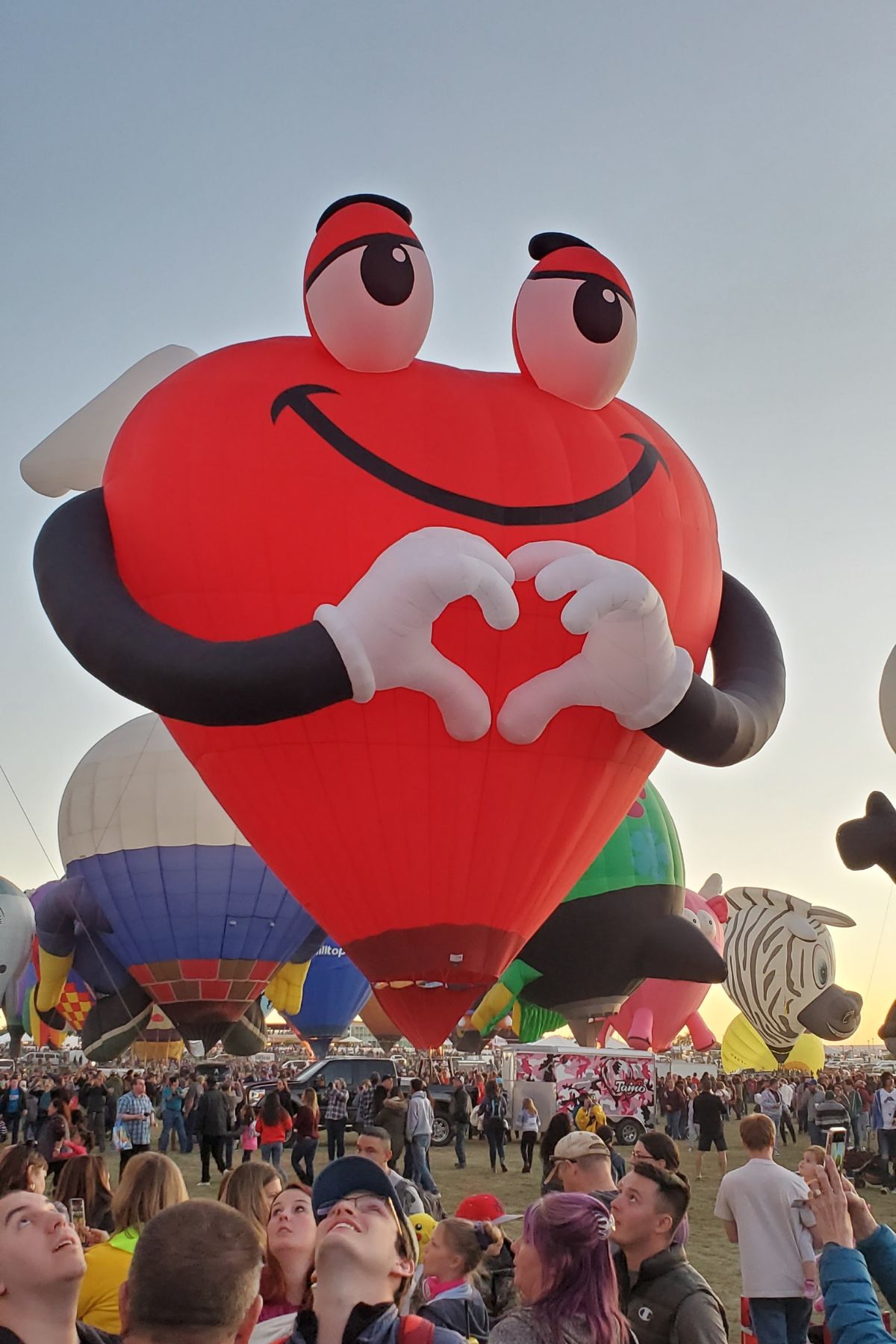 A large red heart-shaped hot air balloon with a smiling face and hands forming a heart rises above a festival crowd, surrounded by colorful balloons—capturing the adventurous spirit of RV Living Full Time.
