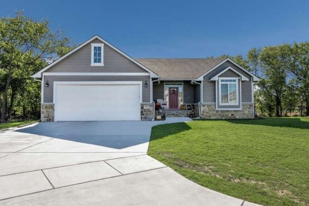 Single-story suburban house with a gray exterior, white trim, double garage, and red front door offers trading stability in a peaceful neighborhood, complemented by a neatly manicured lawn under a clear blue sky.