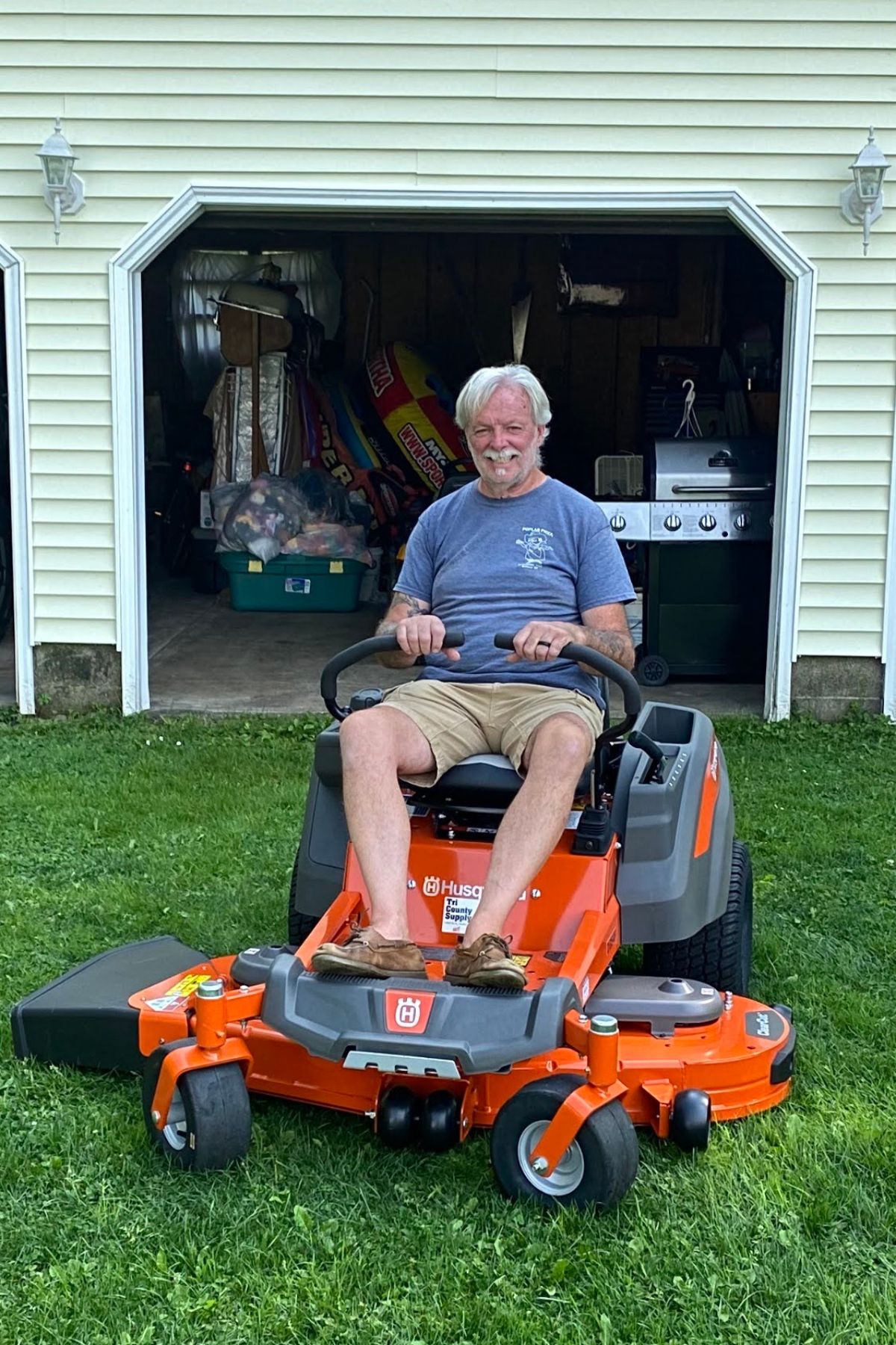 An older man in a blue shirt and shorts sits on an orange Husqvarna riding lawn mower, enjoying the trading stability of routine yard work, in front of an open garage on a grassy lawn.