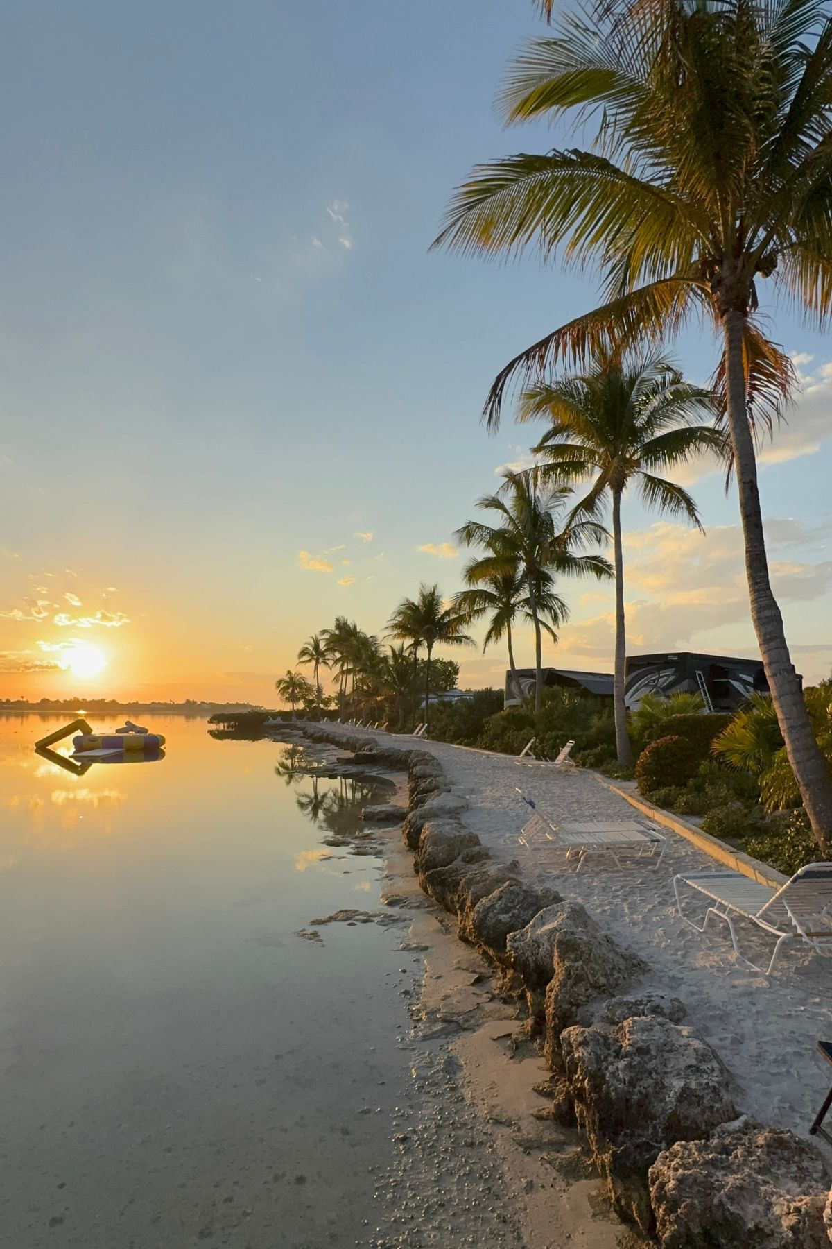 Sunrise over a calm beach with palm trees, lounge chairs, and a rocky shoreline evokes the serene feeling of trading stability, enhanced by a floating platform gently anchored in the tranquil water.