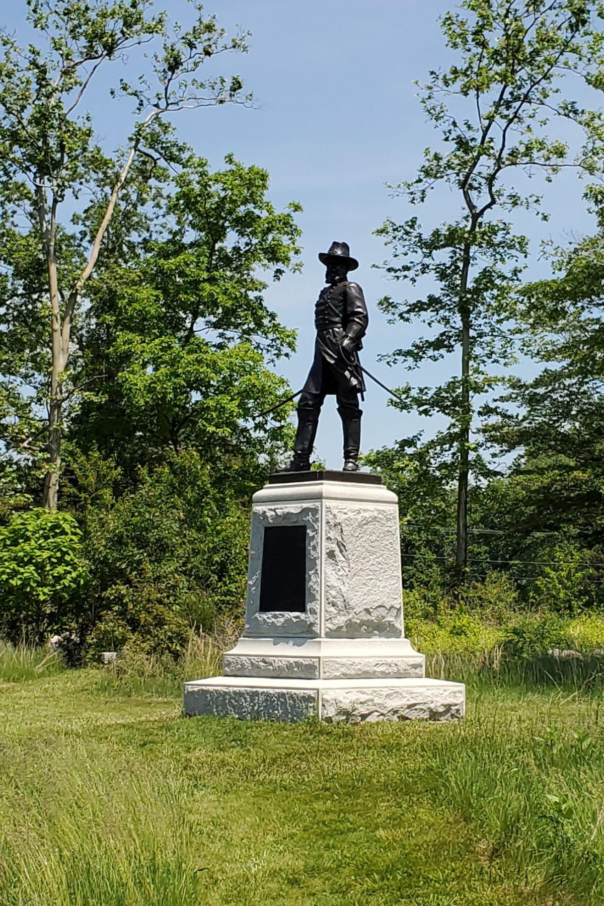 Bronze statue of a man in a hat stands atop a large stone pedestal with a plaque, surrounded by grass and trees under a clear sky, commemorating the Americas 250th Birthday.