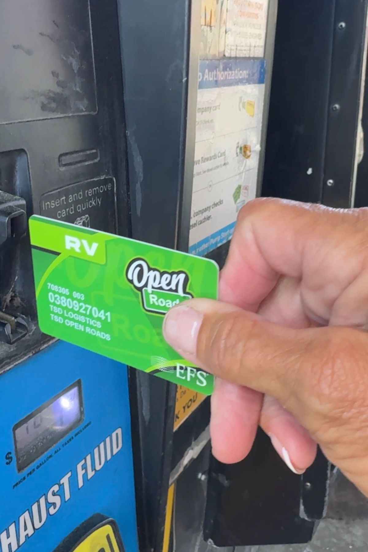 A hand is holding an Open Roads RV card near the card slot of a fuel pump labeled "EXHAUST FLUID," highlighting a convenient way to manage RV fuel costs on your journey.