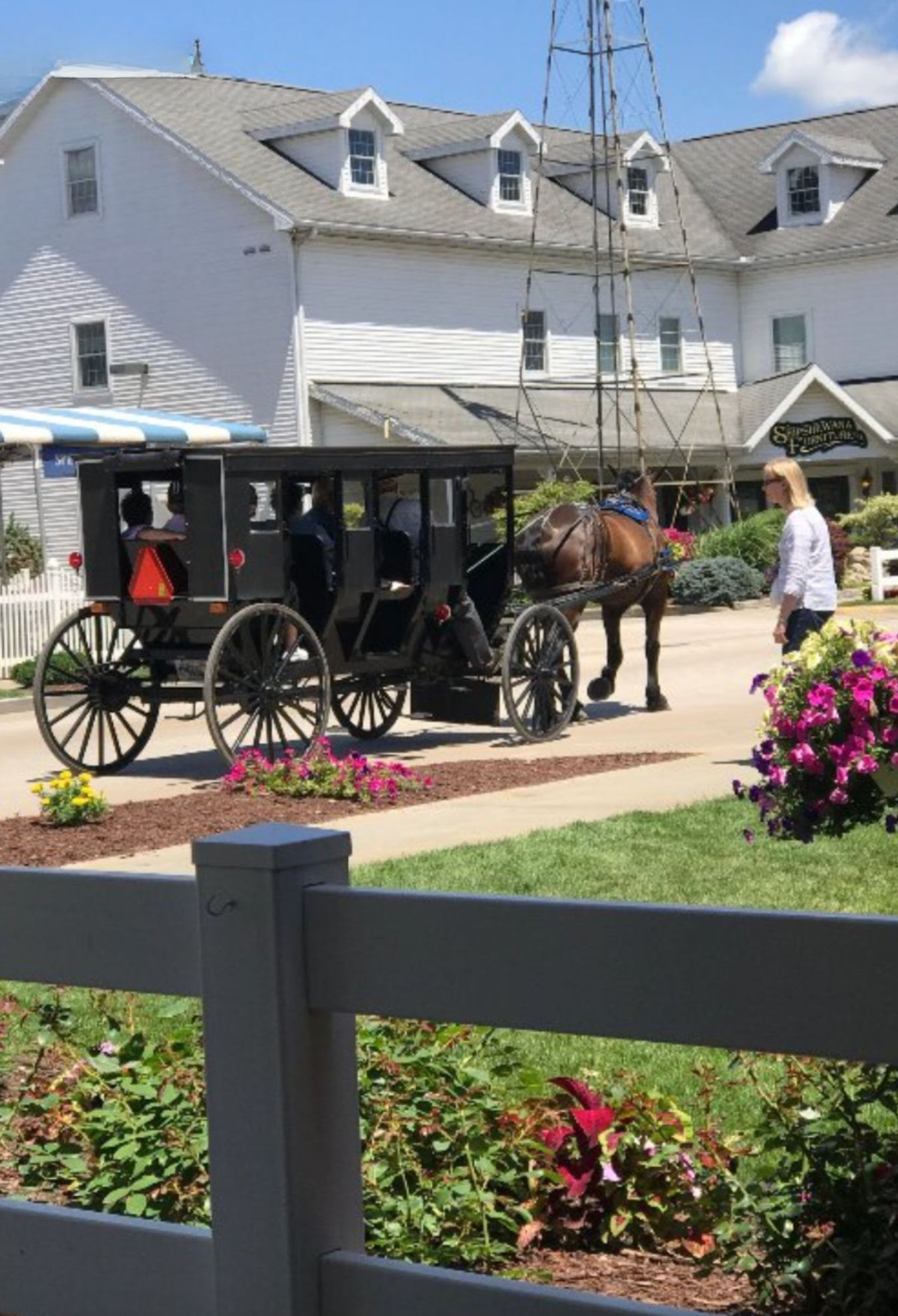 A black horse-drawn carriage is parked outside a large white building, with a person standing nearby and flowers in the foreground, evoking the charm of RV travel across the USA and discovering unique sights along the way.