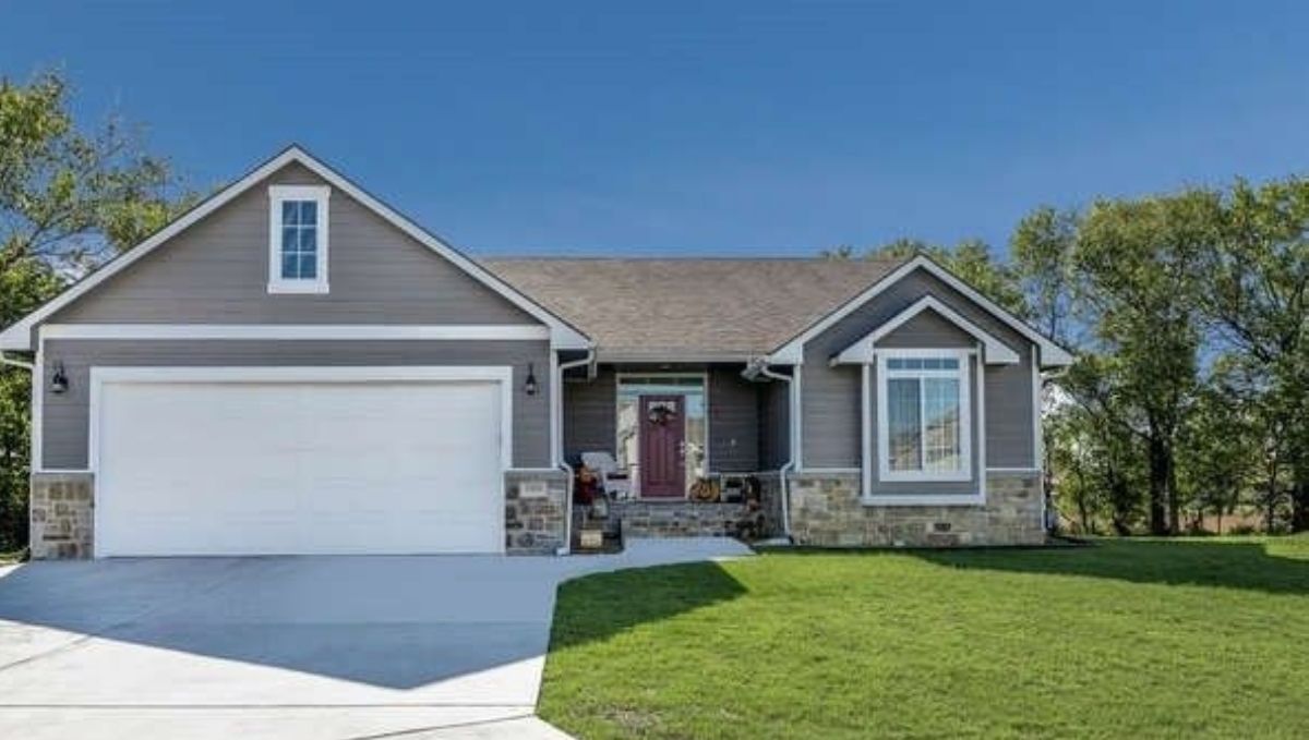 Single-story suburban house with gray siding, white trim, a red front door, attached two-car garage, stone accents, and a well-kept lawn—perfect for those comparing homeownership to Full Time RV Expenses under a clear blue sky.