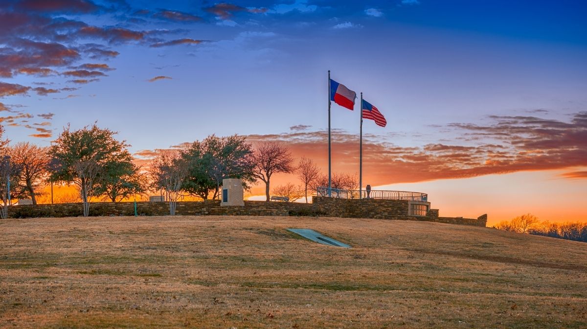 Two flags, one of Texas and one of the United States, stand on flagpoles atop a grassy hill at sunset, with trees and a stone structure in the background—an inspiring scene for America 250th anniversary events.
