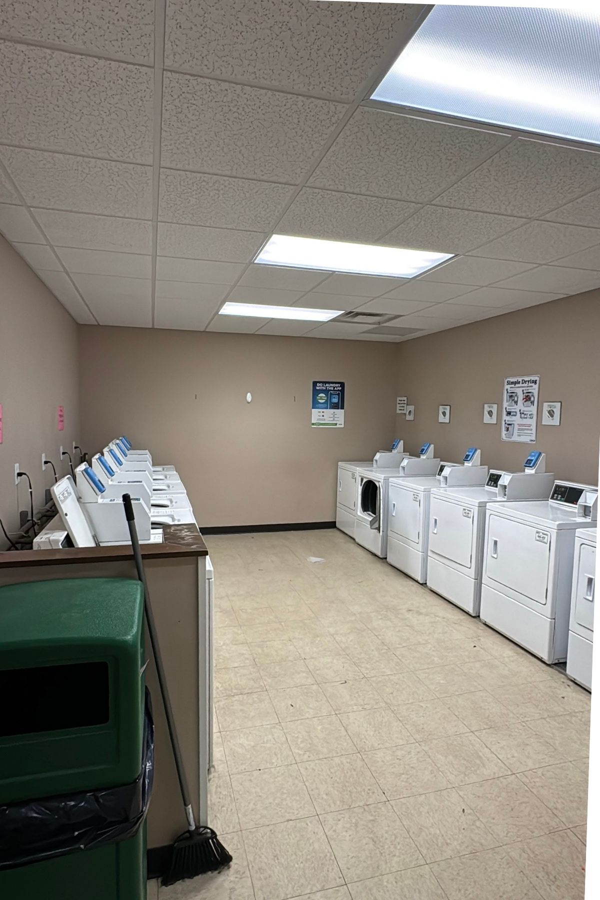 A laundry room at Green Mountain Thousand Trails features several washing machines on the left, dryers on the right, a broom, a green trash bin, and informational signs on the walls.