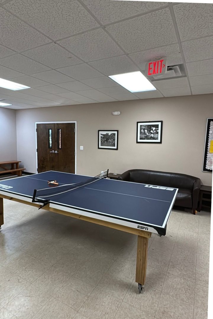 A ping pong table with paddles and a net is set up in a cozy room at Green Mountain Thousand Trails, featuring beige walls, a brown sofa, framed photos, and an exit sign on the ceiling.